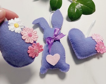 May include: Three lavender felt Easter ornaments: an egg shape with white and pink flowers, a bunny with a purple bow and pink heart, and a heart with pink flowers. White ribbons are attached for hanging.