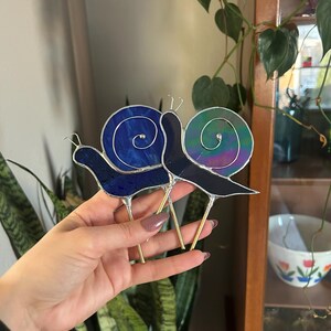 May include: Two stained glass snail figurines with gold metal stems. The snail on the left has a blue shell with a spiral design. The snail on the right has a green, iridescent shell.