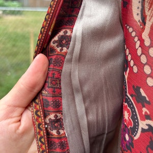 May include: Close-up of the inside of a red and gold patterned garment. The fabric is lined with a smooth, silver-colored material.