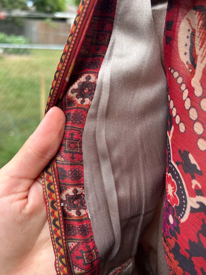 May include: Close-up of the inside of a red and gold patterned garment. The fabric is lined with a smooth, silver-colored material.