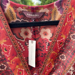 May include: A red, orange, and yellow patterned dress with a V-neckline. The dress has a tag that says "Anthropologie" and a label that says "Ranna Gill".