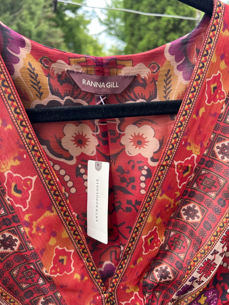 May include: A red, orange, and yellow patterned dress with a V-neckline. The dress has a tag that says "Anthropologie" and a label that says "Ranna Gill".