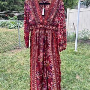 May include: A long, red, and purple patterned maxi dress with long sleeves and a V-neckline. The dress has a fitted bodice and a flowing skirt. The fabric is sheer and lightweight.