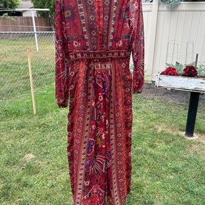May include: A long-sleeved maxi dress with a red, brown, and purple paisley print. The dress has a V-neckline and a fitted bodice. The skirt is flowy and has a slight A-line shape.