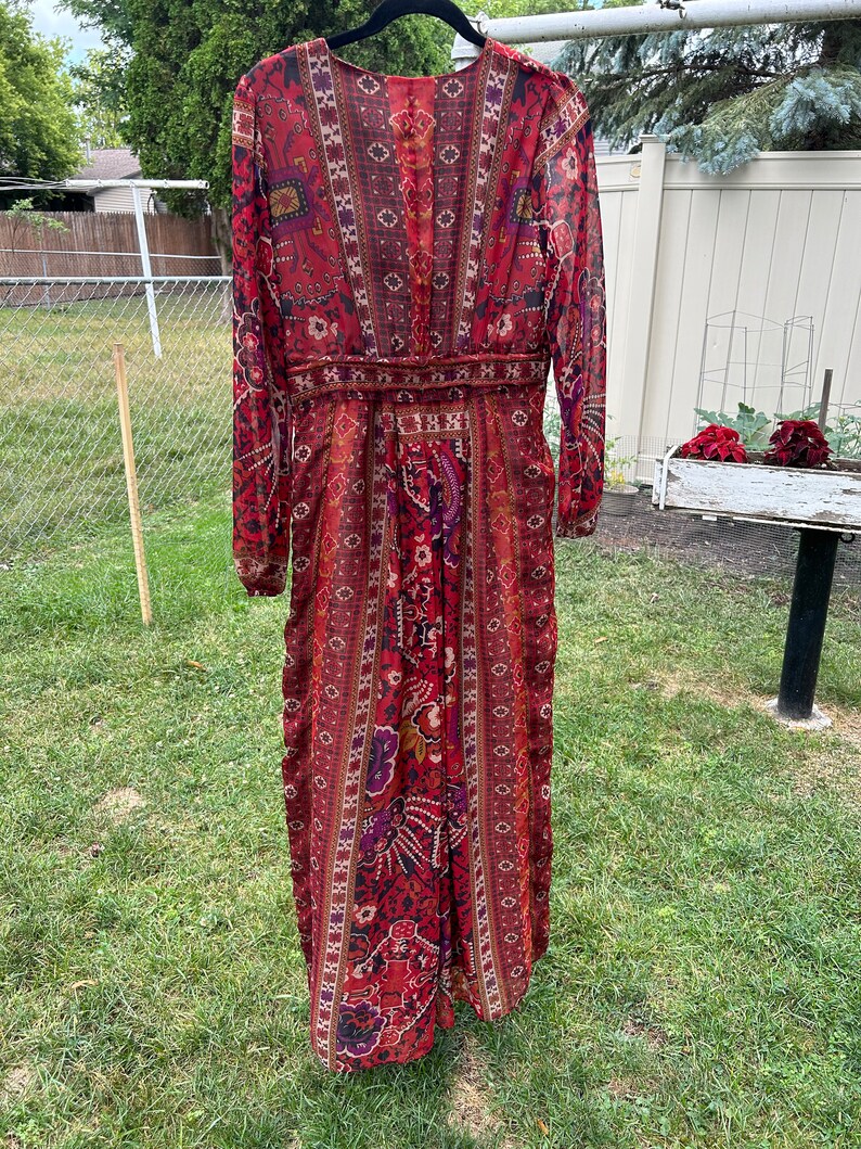 May include: A long-sleeved maxi dress with a red, brown, and purple paisley print. The dress has a V-neckline and a fitted bodice. The skirt is flowy and has a slight A-line shape.