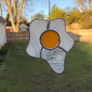 May include: A stained glass suncatcher in the shape of a fried egg. The egg features a bright yellow yolk, surrounded by a white, textured egg white. A silver chain is attached for hanging. The suncatcher is displayed outdoors.