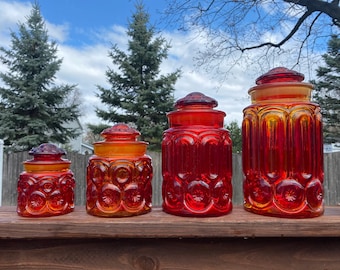 May include: Four vintage glass canisters in varying sizes, featuring a vibrant orange and red color scheme. Each jar has a matching lid and a decorative pattern of circles and arches. The jars are arranged in a row, showcasing their different heights.