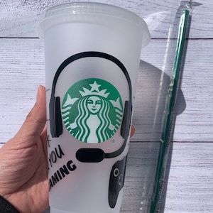May include: A white reusable cup with a black and green Starbucks logo, black headphones, and a black video game controller. The text "Let You Gaming" is written on the cup.