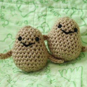 May include: Two brown crocheted potato toys with black eyes and smiling mouths. The potatoes are sitting on a green fabric with white writing.