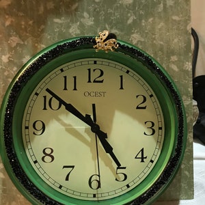 May include: Green wall clock with black accents and a decorative gold-colored embellishment at the top. The clock face is white with black numerals and hands. The brand name "OCEST" is printed on the clock face.