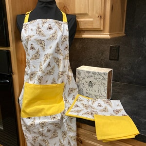 May include: A white apron with a yellow pocket and straps, featuring a floral and bee pattern. A matching oven mitt, pot holder, and a folded yellow towel are also visible. A decorative box with the word "KIND" is also present.