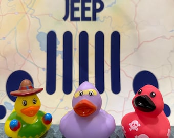 May include: Three rubber ducks with unique designs. One yellow duck wears a sombrero and a green shirt, another is purple with a mask, and the third is red with skull and crossbones. The background features the text "DUCKDUCK JEEP".