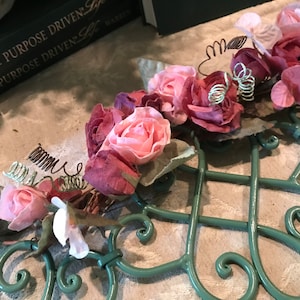 May include: A decorative metal trellis painted in a soft green, adorned with clusters of pink and burgundy artificial roses and delicate wire accents. The trellis is placed on a neutral-toned surface.