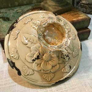 May include: A cream-colored, decorative lid with a clear glass knob. The lid features embossed floral and leaf designs with distressed black accents. The knob has a textured, amber-colored interior. The lid appears to be made of metal.
