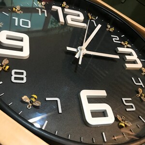 May include: Black wall clock with white numerals and hands. The clock features decorative gold-colored bee accents around the face. The clock has a black frame and is likely for home decor.