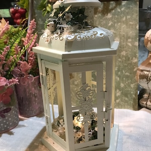 May include: A white decorative lantern with a hinged door and a decorative top. The lantern features a small bird and floral accents. The interior is illuminated, and the lantern sits on a white base. The lantern is a decorative home accent.