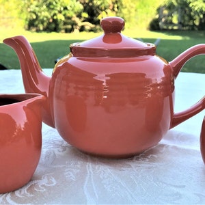 May include: A terracotta colored teapot, creamer, and sugar bowl set. The teapot has a round body and a curved spout. The creamer and sugar bowl are both round and have lids.