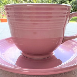 May include: A pink teacup and saucer set. The cup has a wide, rounded shape and a handle. The saucer is a simple, round shape.