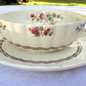 May include: Cream-colored soup bowl with two handles, resting on a matching saucer. The bowl and saucer feature a floral pattern with red, pink, and purple flowers. The edges of the saucer have a scalloped design.
