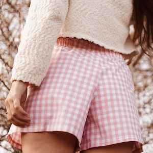 May include: Pink and white gingham shorts featuring an elasticized waistband. The shorts are paired with a textured cream-colored long-sleeve top. The fabric pattern consists of a classic checkered design in a soft pastel pink and white color scheme.