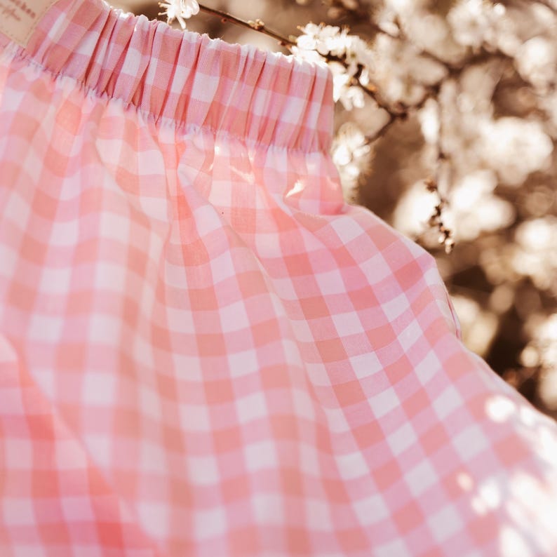 May include: A pair of shorts featuring a light pink and white gingham check pattern. The garment has an elasticized waistband with gathered fabric detailing, creating a soft, pleated effect across the top edge of the checkered textile.