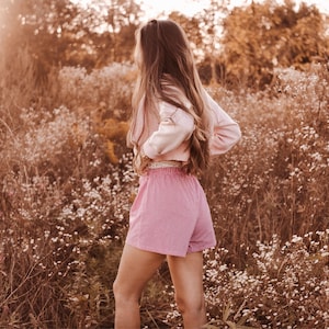 May include: A light pink long-sleeved cardigan paired with pink and white gingham shorts featuring an elastic waistband. The shorts have a classic checkered pattern and a relaxed fit, while the cardigan is worn open over the waistline.