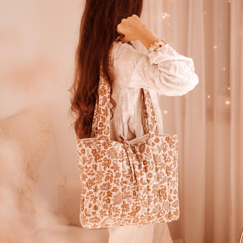 May include: A beige tote bag with a floral pattern in shades of brown and cream. The bag has a quilted appearance and a drawstring closure. The straps are made of the same patterned fabric. The bag is being held by a person.