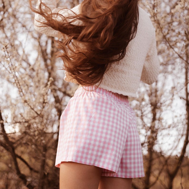 May include: A pair of light pink and white gingham shorts with an elastic waistband, paired with a textured cream-colored long-sleeve top. The shorts feature a classic checkered pattern and a relaxed fit, while the top has a soft, woven appearance.