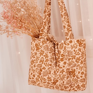 May include: A beige tote bag with a brown floral pattern, filled with dried pink flowers. The bag has two handles and a knotted closure. The floral design includes leaves and various flower shapes.