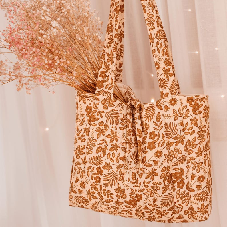 May include: A beige tote bag with a brown floral pattern, filled with dried pink flowers. The bag has two handles and a knotted closure. The floral design includes leaves and various flower shapes.