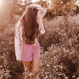 May include: A light pink long-sleeved knit cardigan worn over a matching light pink top, paired with red and white gingham patterned shorts featuring an elasticized waistband, captured in a soft, warm-toned outdoor setting.