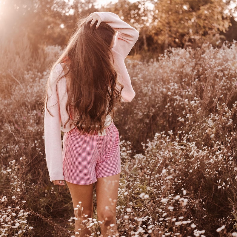 May include: A light pink long-sleeved knit cardigan worn over a matching light pink top, paired with red and white gingham patterned shorts featuring an elasticized waistband, captured in a soft, warm-toned outdoor setting.