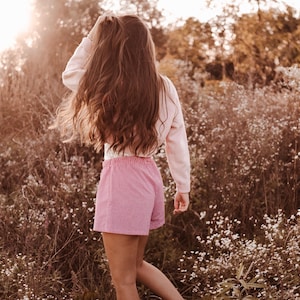 May include: A long-sleeved light pink pullover sweater paired with red and white gingham patterned shorts featuring an elasticized waistband, worn by an individual with long brown hair standing in a field of small white wildflowers.