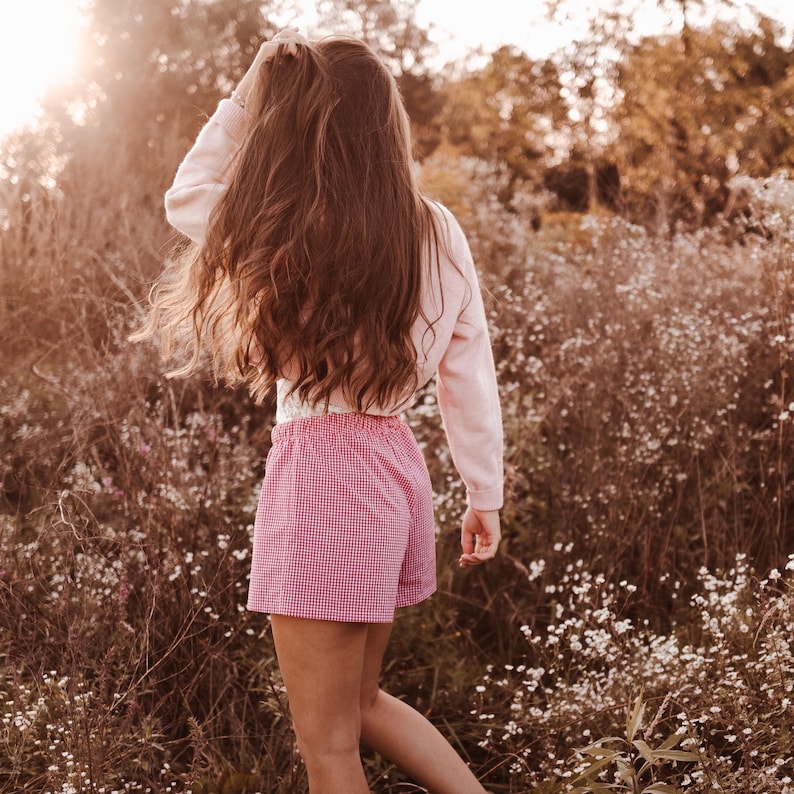 May include: A long-sleeved light pink pullover sweater paired with red and white gingham patterned shorts featuring an elasticized waistband, worn by an individual with long brown hair standing in a field of small white wildflowers.