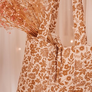 May include: A beige tote bag with a brown floral pattern, filled with dried pink flowers. The bag has two handles and a knotted closure. The floral design includes leaves and flowers.