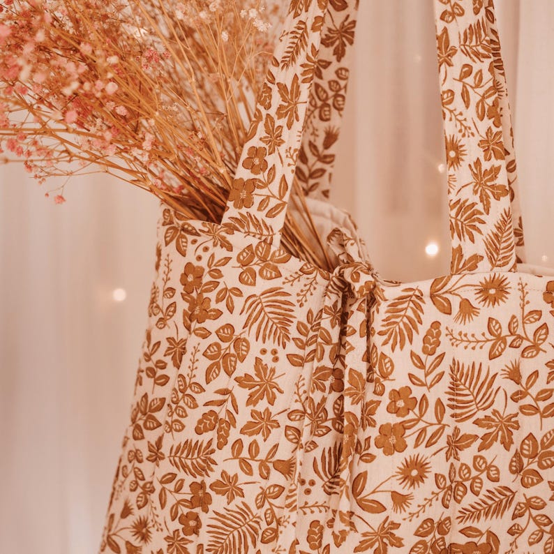 May include: A beige tote bag with a brown floral pattern, filled with dried pink flowers. The bag has two handles and a knotted closure. The floral design includes leaves and flowers.