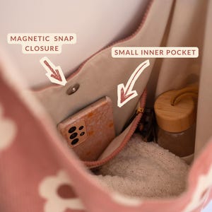 May include: Pink tote bag with a white floral pattern. The interior features a small inner pocket and a magnetic snap closure. A smartphone, a water bottle, and a folded sweater are visible inside the bag.