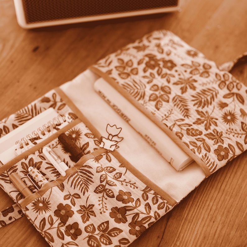May include: A fabric organiser with a floral pattern, open to reveal a notebook, pens, and a book. The fabric is brown and cream with a floral design. The organiser has multiple pockets and compartments.