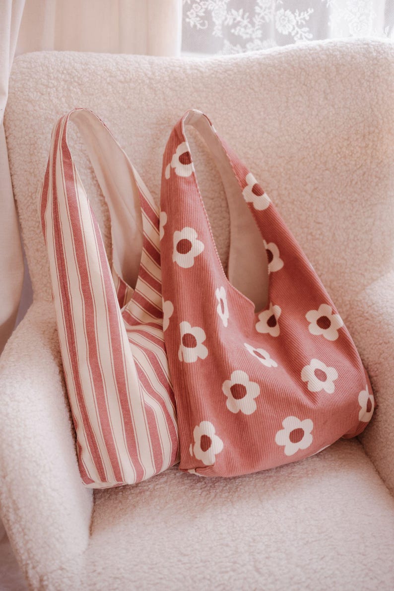 May include: Two pink tote bags. One has a vertical stripe pattern in white and pink. The other is a solid pink colour with a white daisy pattern. Both bags have a rounded shape and a single shoulder strap.
