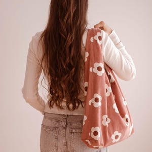 May include: A dusty pink corduroy tote bag with a white floral pattern is carried over the shoulder. The bag is a soft, unstructured style. The person in the image is wearing a white long-sleeve shirt and light wash jeans.