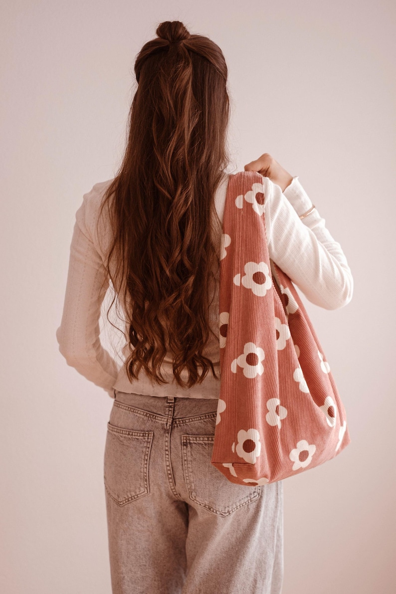 May include: A dusty pink corduroy tote bag with a white floral pattern is carried over the shoulder. The bag is a soft, unstructured style. The person in the image is wearing a white long-sleeve shirt and light wash jeans.