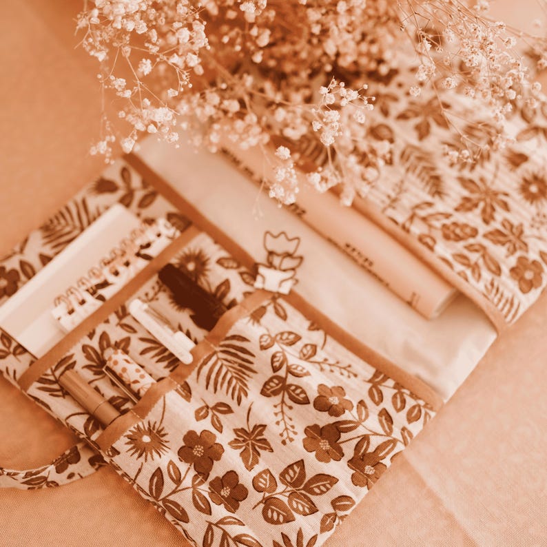 May include: A beige pencil case with a brown floral pattern, holding pens, pencils, and a small notebook. The case is open, revealing its contents. A sprig of white flowers is placed on top, creating a soft, natural aesthetic.