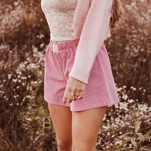 May include: Red and white gingham shorts with an elastic waistband and a small rectangular label on the front. The shorts are paired with a white lace top and a light pink long-sleeved cardigan, creating a layered outfit with a classic pattern.