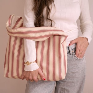 May include: A striped tote bag in red and cream. The bag has a wide opening and a folded-over top. The bag is made of a sturdy fabric. The bag is held by a person wearing a white long-sleeved shirt and grey jeans. The bag is suitable for shopping.