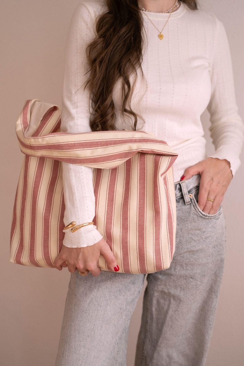 May include: A striped tote bag in red and cream. The bag has a wide opening and a folded-over top. The bag is made of a sturdy fabric. The bag is held by a person wearing a white long-sleeved shirt and grey jeans. The bag is suitable for shopping.