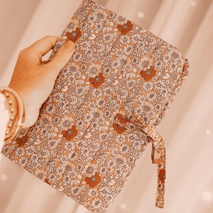 May include: A floral patterned journal with a tie closure. The journal features a repeating pattern of white and rust-coloured flowers on a tan background. The journal is held in a hand, with a gold bracelet visible.