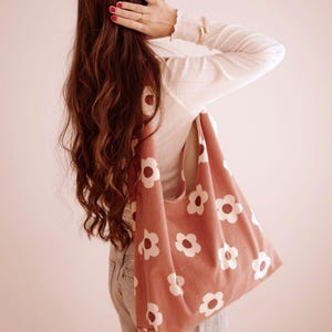 May include: A coral corduroy tote bag featuring a white daisy print. The bag is carried over the shoulder. The background is a light pink. The bag is a stylish accessory for carrying items. The bag is a fashionable item.
