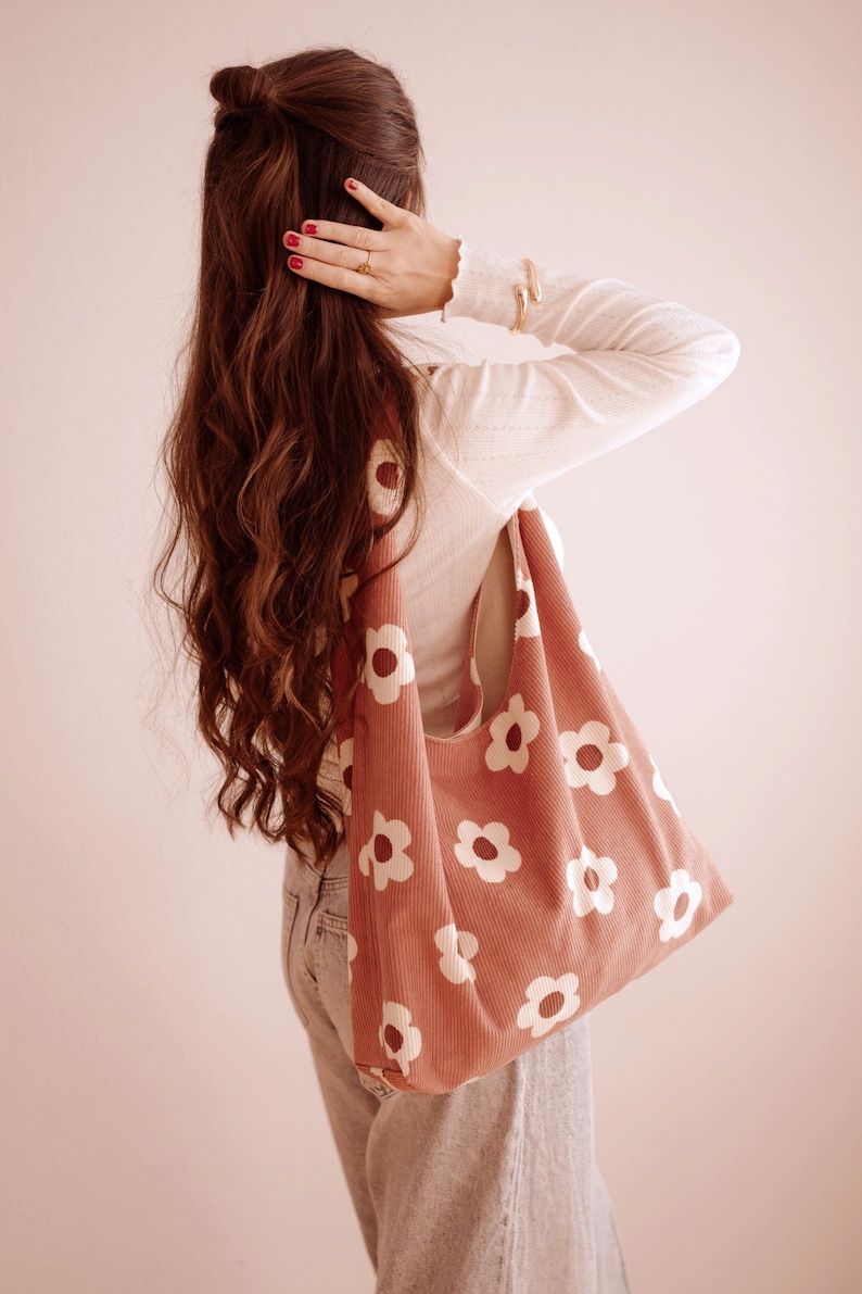 May include: A coral corduroy tote bag featuring a white daisy print. The bag is carried over the shoulder. The background is a light pink. The bag is a stylish accessory for carrying items. The bag is a fashionable item.