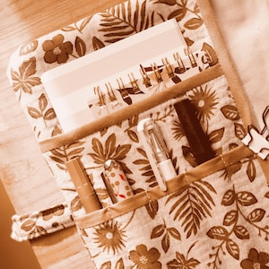 May include: A beige notebook cover with a brown floral pattern, holding a spiral-bound notebook and pens. The cover has a pocket containing pens and a lipstick. The notebook is white with a metal spiral binding. The design features leaves and flowers.