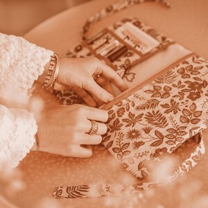 May include: A floral patterned pencil case in shades of brown and cream, open to reveal pencils and a small pocket. The case has a matching strap and is being held open.